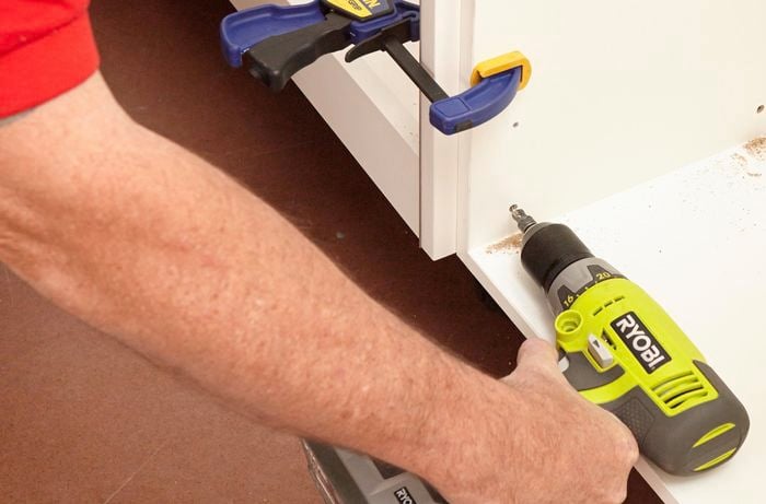 Two kitchen cabinets being screwed together with a power drill, while held together with clamps