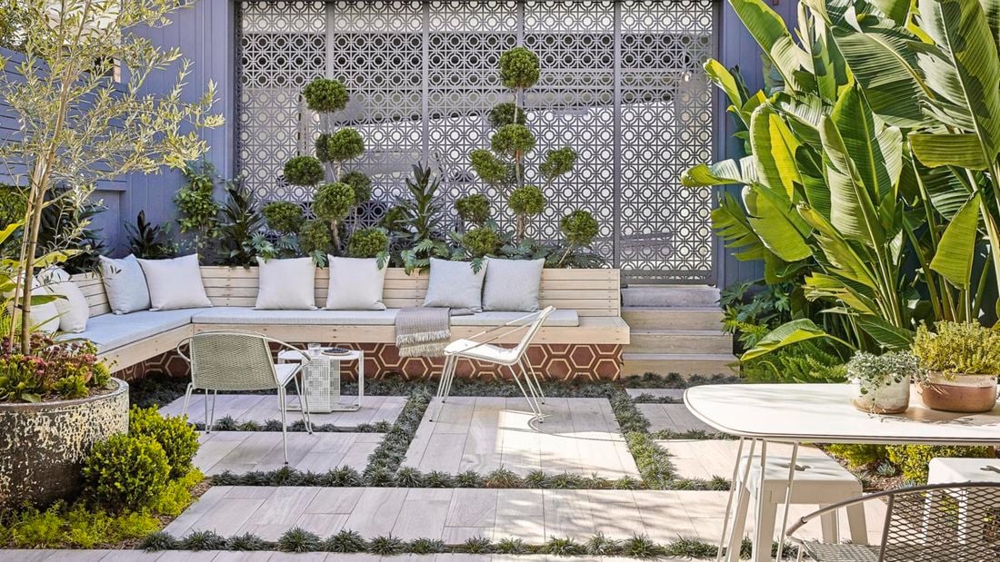 An outdoor room with timber bench seats, concrete pavers, metal wall screens and green plants
