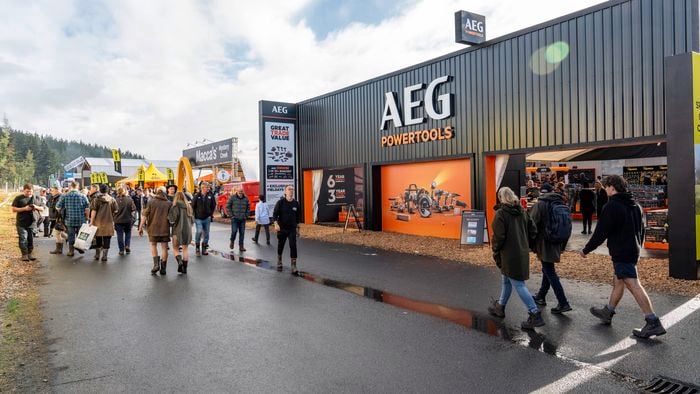 Crowd of people walking along a wet pathway at an outdoor trade event, passing a large black and orange AEG Powertools exhibition stand, with branded signage, display tents, and other vendor booths visible under a partly cloudy sky.