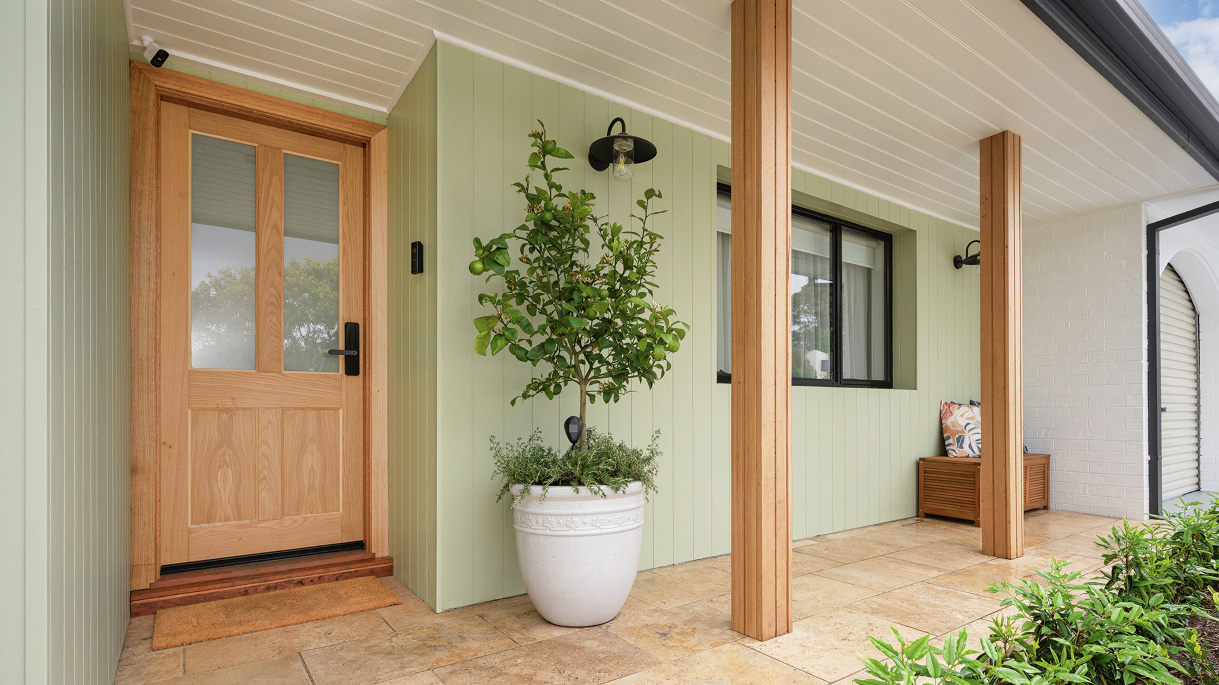 A light green house exterior with a wooden front door, featuring a security camera, video doorbell, digital door lock, and a porch light, alongside a potted plant on a tiled porch.