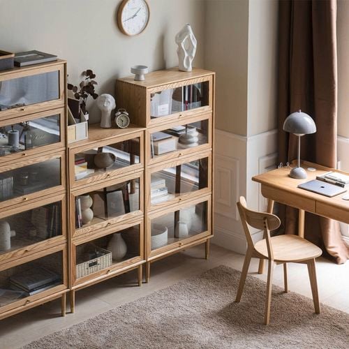 A warm, modern workspace featuring natural wood bookshelves with glass doors and a minimalist writing desk.