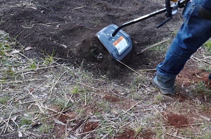 A person turning over soil using a tiller attachment on a brush cutter