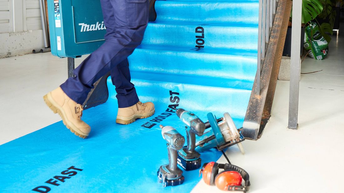 Construction site interior showing protective floor covering installed over stairs and flooring, with power tools and hearing protection placed nearby and a person carrying a tool case up the stairs.