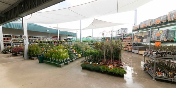 Outdoor Bunnings Garden Centre featuring rows of potted plants, shrubs and young trees arranged beneath large white shade sails. Garden products and landscaping materials are displayed on surrounding racks, with wide walkways and covered retail structures in the background.