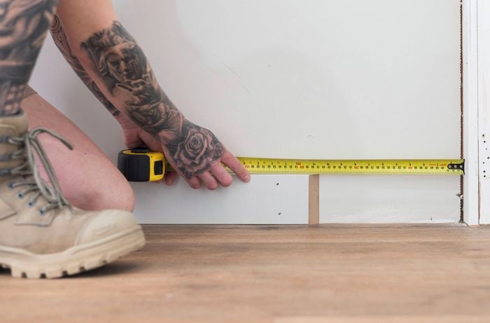 A person with tattooed arms is measuring the length of a wall where the skirting boards installation will be done.