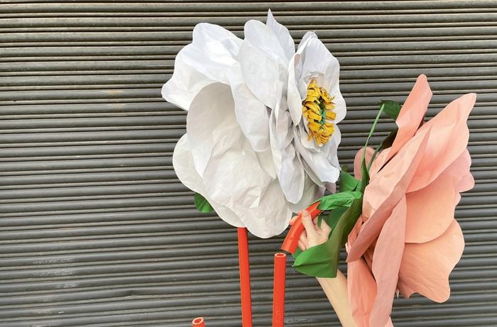 A hand slotting a giant paper flower attached to a conduit sweep bend to an orange conduit, next to a white flower, both with green leaves.