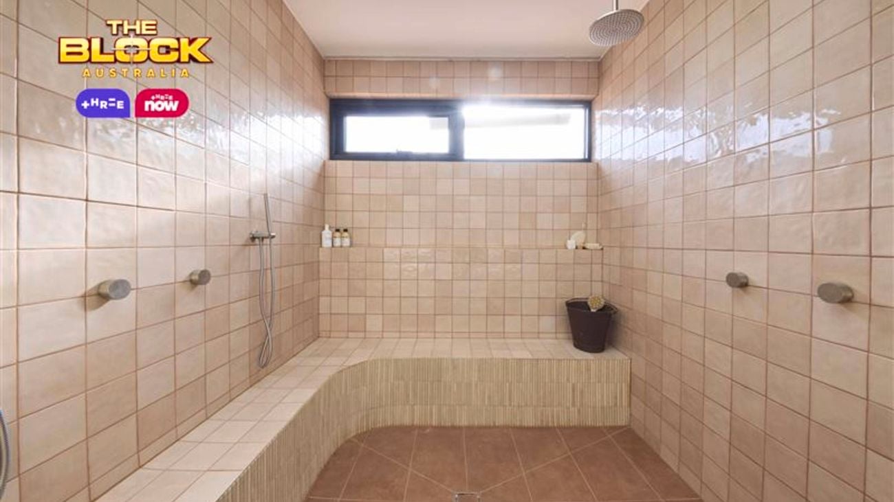 Spacious shower room with pink tiled walls, brown tiled floor, built-in bench, multiple showerheads, and natural light from a high window. 