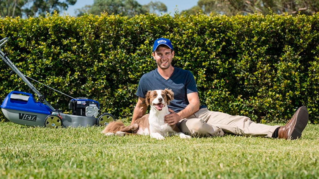 A man with his dog, and a mower, sit on the backyard in the sun.
