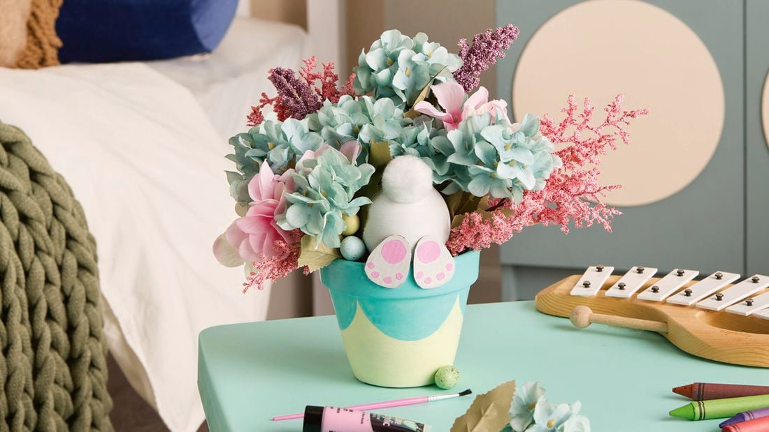 A painted plant pot decorated with an Easter theme is filled with artificial flowers and a small bunny figure, sitting on a craft table in a bedroom setting.