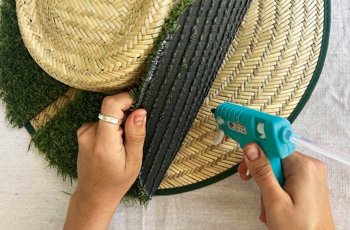 A hand holds a piece of artificial turf as it is attached to the brim of a Bunnings straw hat using a hot glue gun.