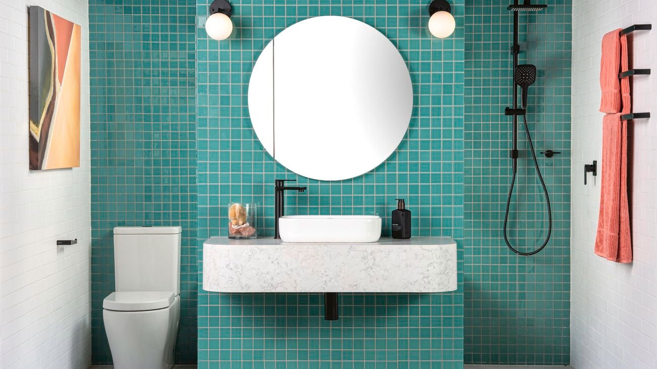 A bathroom with green mosaic tiled walls. A toilet and shower are either side of a floating marble countertop with a large round mirror above it.