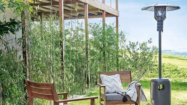 Outdoor living space with patio heater. The area is surrounded by lush green grass and what appears to be bamboo plants in the background. A pair of chairs are positioned by a fire at the centre with a dog in the foreground.