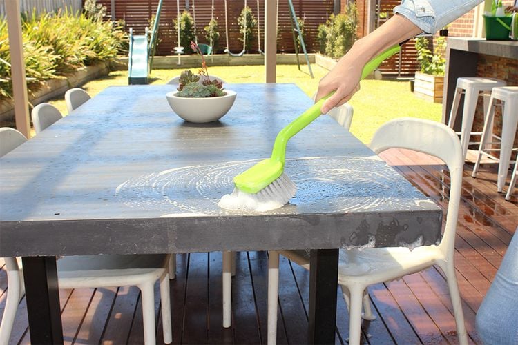 Person cleaning an outdoor table with a long-handled scrubbing brush