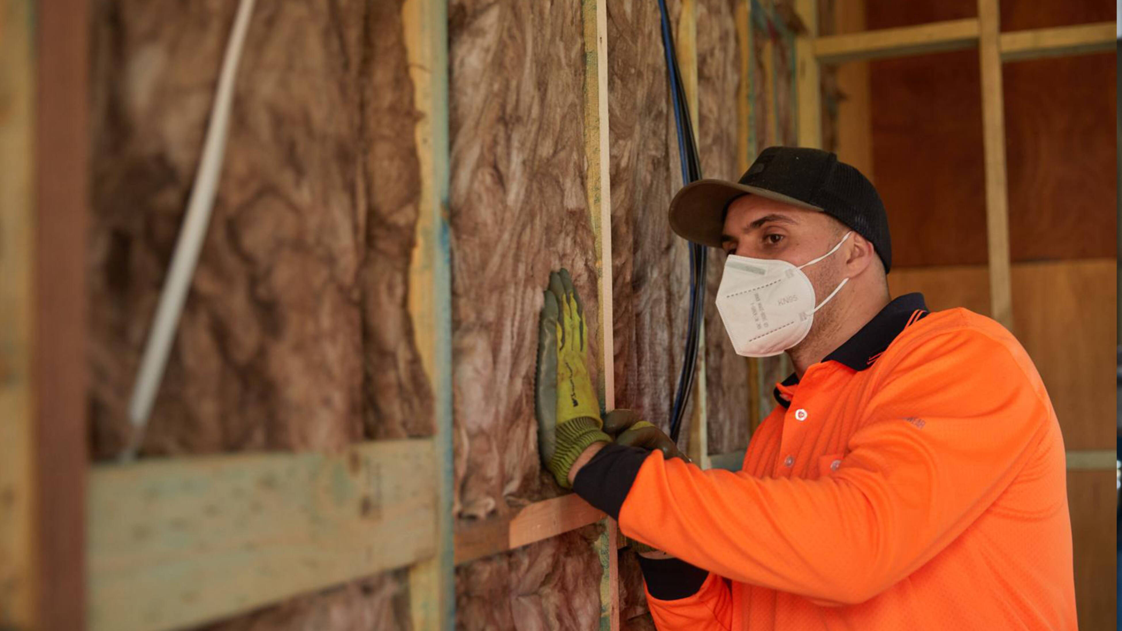 A worker in an orange shirt installing insulation material into a timber wall frame. A tradesperson leaning against the back of an open van with tools and equipment visible inside, alongside a dog sitting inside the van.