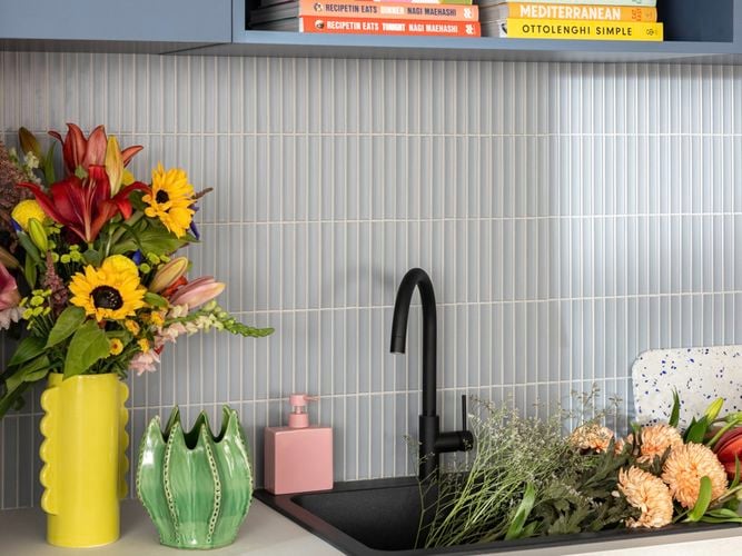 Blue modern kitchen with white benchtop with crockery on it, a light blue splashback and a sink with flowers in it.