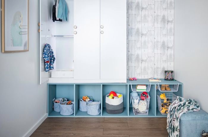 A child's room with a wardrobe on a shelving unit against a wall and a toy box with a blanket on it.
