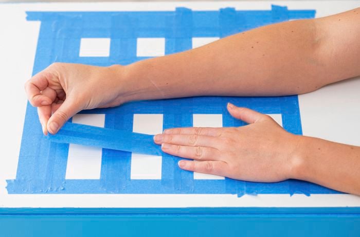 A person is applying painter’s tape to create a grid pattern on a painted toy box lid.