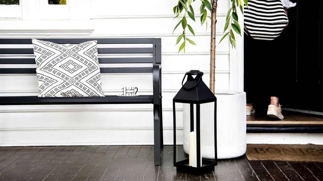 Front porch with a black bench and patterned cushion beside a tall potted tree. A large lantern with a candle on the floor, a wall light, and a striped bag hanging near the black front door.