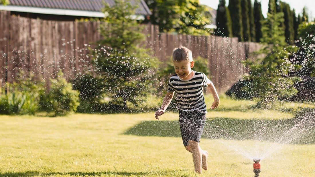 a child in a black and white t-shirt, running through a sprinkler and enjoying summer water play outdoors