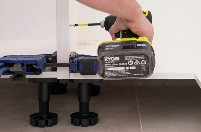 A person is using a power drill to join two cabinet units, secured in place with clamps and supported by adjustable legs