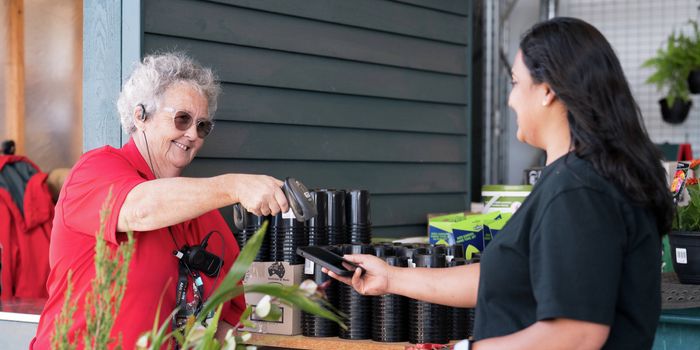 A Bunnings Team Member holding a scanner above a mobile phone a lady is holding in the outdoor nursery of a Bunnings store.