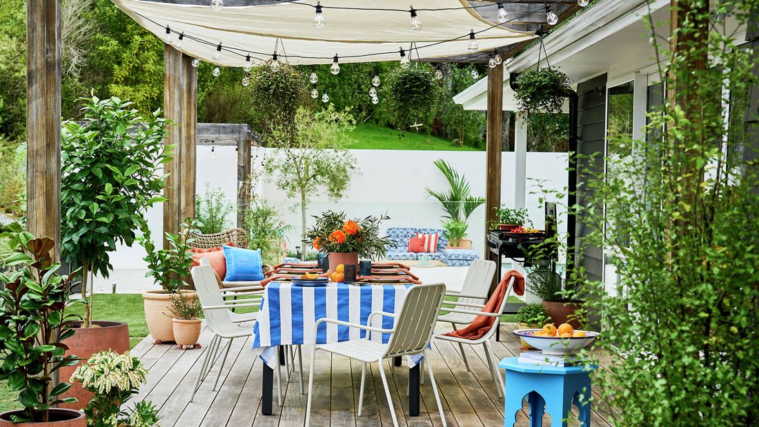 Outdoor dining area on a timber deck with a blue and white striped tablecloth, surrounded by potted plants and chairs, with a shade sail overhead