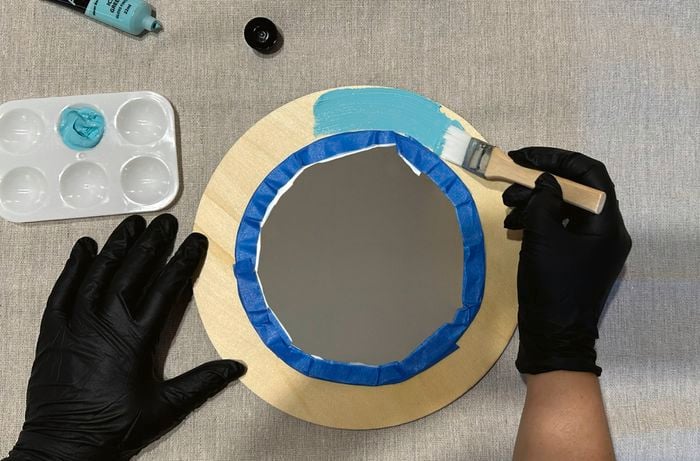 Hands wearing black gloves painting the wooden frame of a round mirror with teal acrylic paint, with the mirror masked off using blue painter’s tape.