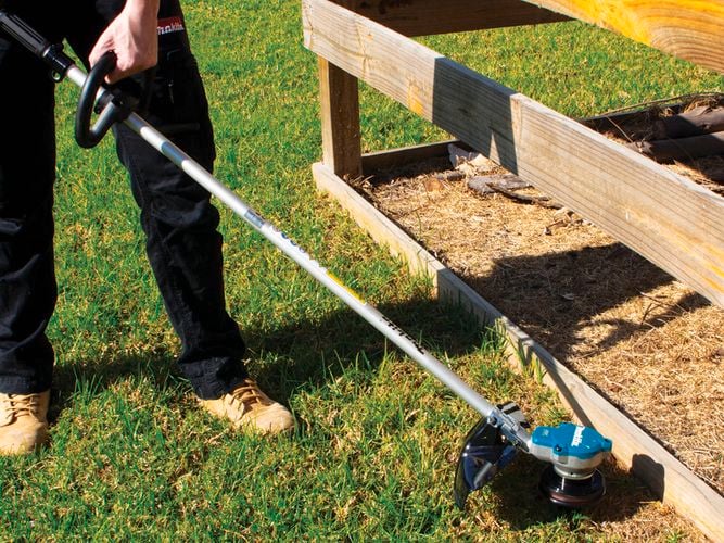 Person trimming grass near a wooden fence using a string trimmer, with green grass and a patch of bare soil nearby.