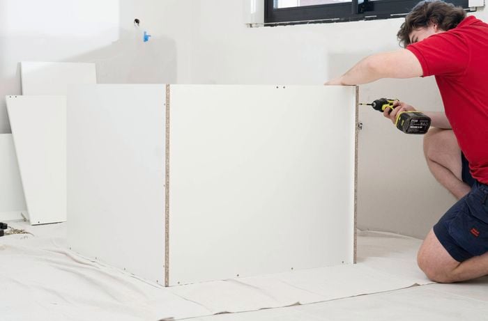 A person in a red t-shirt is assembling the side panels of a corner base cabinet using a power drill.