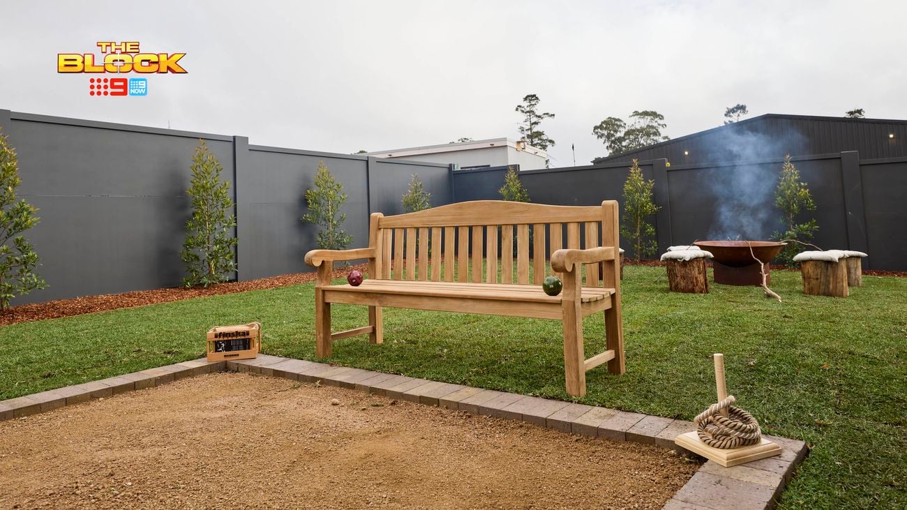 A backyard, with an outdoor chair, firepit with stumps of wood in front of it and trees in it.