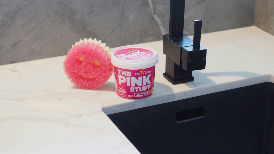 A pink Scrub Mommy sponge and a tub of The Pink Stuff cleaning paste sitting on a benchtop beside a black tap.
