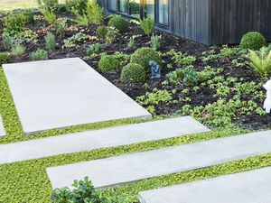A concrete stepping-stone path with green groundcover between slabs beside a mulched garden of small shrubs, ornaments and round hedges.