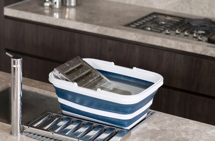 The image shows a collapsible bucket with sub merged oven trays and range hood filters, next to an over-sink drying rack on a kitchen counter.