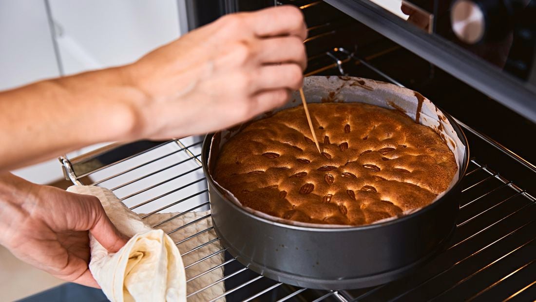 A cake being baked in a cake tin, sitting in an open oven, with the cook piercing the top with a skewer