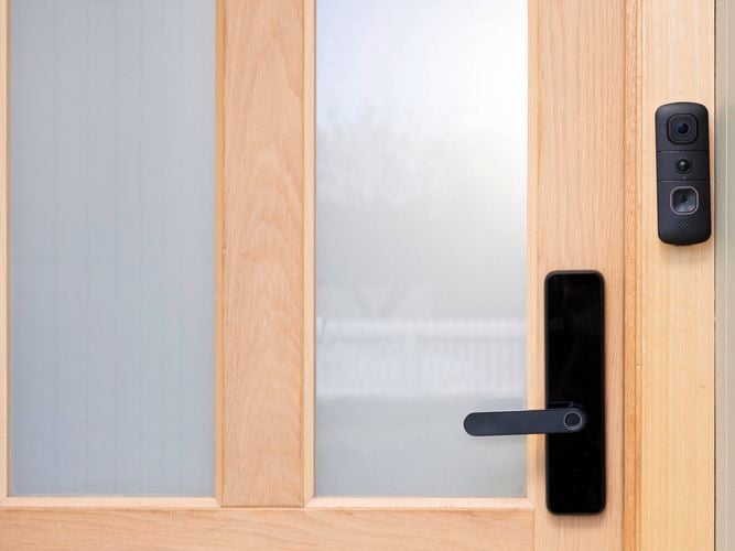 A black Swann video doorbell is mounted beside the handle of a wooden front door with frosted glass panels. 
