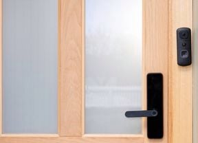 A black Swann video doorbell is mounted beside the handle of a wooden front door with frosted glass panels. 