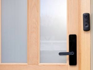 A black Swann video doorbell is mounted beside the handle of a wooden front door with frosted glass panels. 