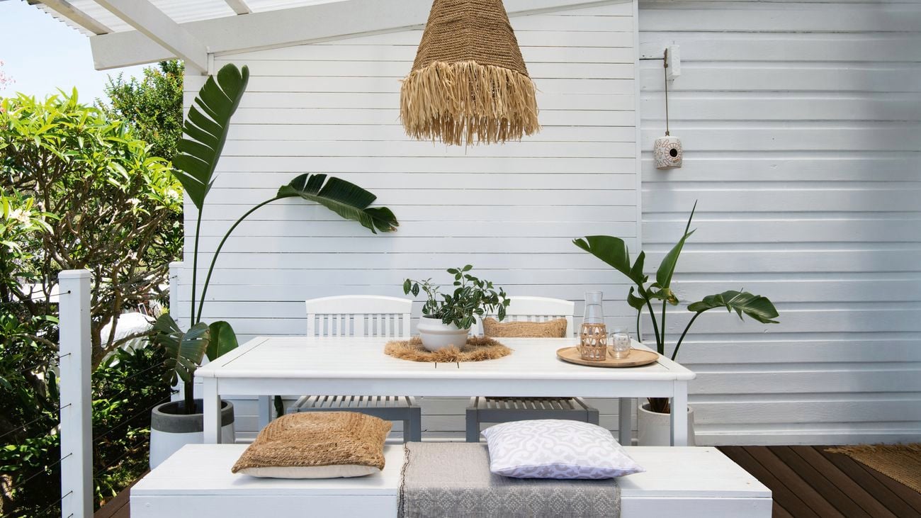 A painted white timber bench and table contrasts against lush green potted plants. A jute lamp shade with fringe hangs above.