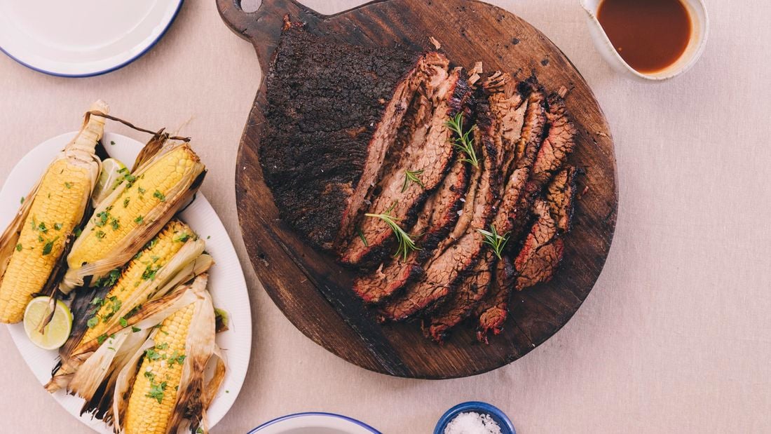 A perfectly cooked brisket on a table with some gravy and char grilled corn cobs