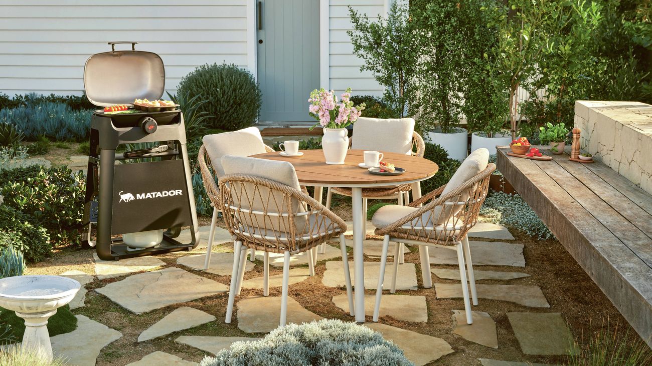 Outdoor dining set with a round wooden table, woven chairs, and pink flowers on a patio
