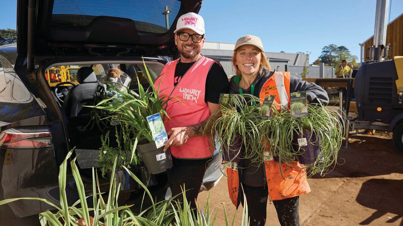Two adults in hi-vis clothing stand beside a hatchback with its boot open, each holding several potted plants. The car boot is filled with plants, with construction equipment and a worker visible in the background.