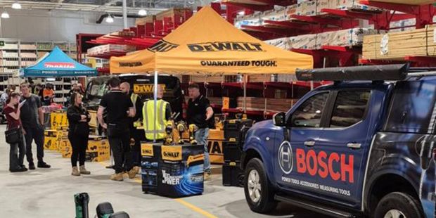 Indoor hardware store event with brand displays and tools; a Bosch-branded utility vehicle is parked on the right, DeWalt canopy tents and product stands are set up in the middle, and several people are gathered around demonstrations. Shelving with timber and materials lines the background, while power tools are displayed on a table in the foreground.