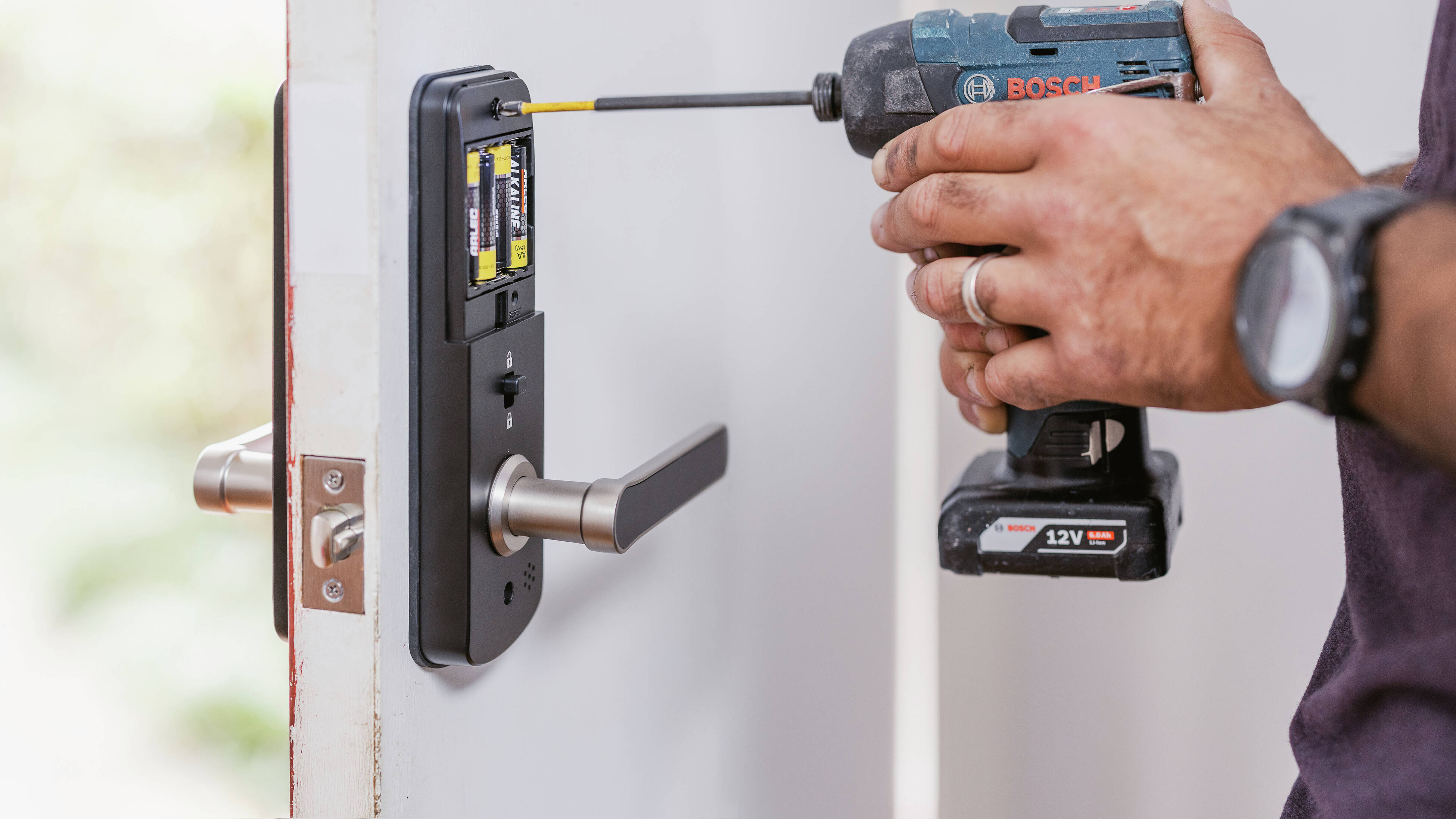 “Person using a power drill to install a smart door lock on a white door, with batteries visible in the lock housing.”