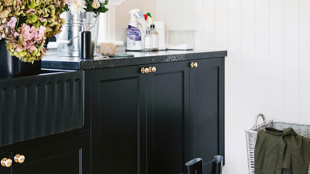 A laundry room with black cabinetry, a black sink, a black countertop with flowers in a bucket, and a pair of black gumboots on a light-coloured tiled floor.