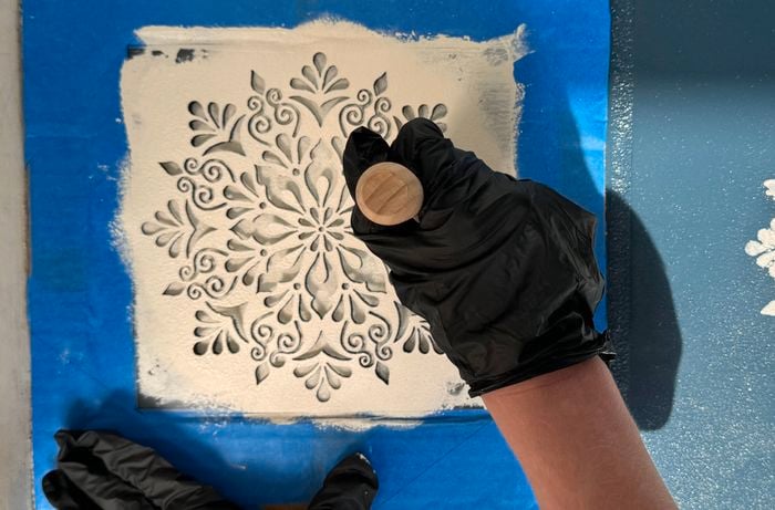 A stencil taped to a tile is being painted by gloved hands using a round brush