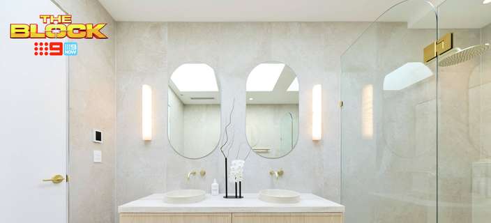 Modern bathroom with a floating timber double vanity, white basins, oval mirrors, wall lights, brass accents and a glass-enclosed shower. “The Block” logo appears in the upper-left corner.