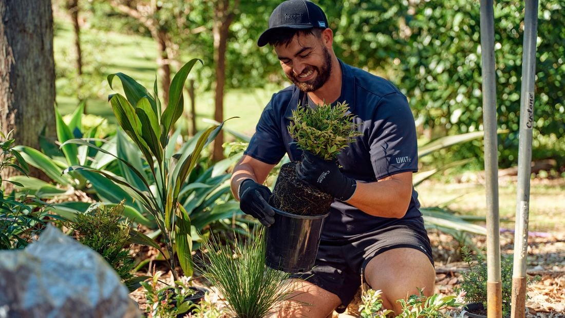 Person kneeling outdoors placing a small potted plant into soil among garden plants, tools, and greenery.