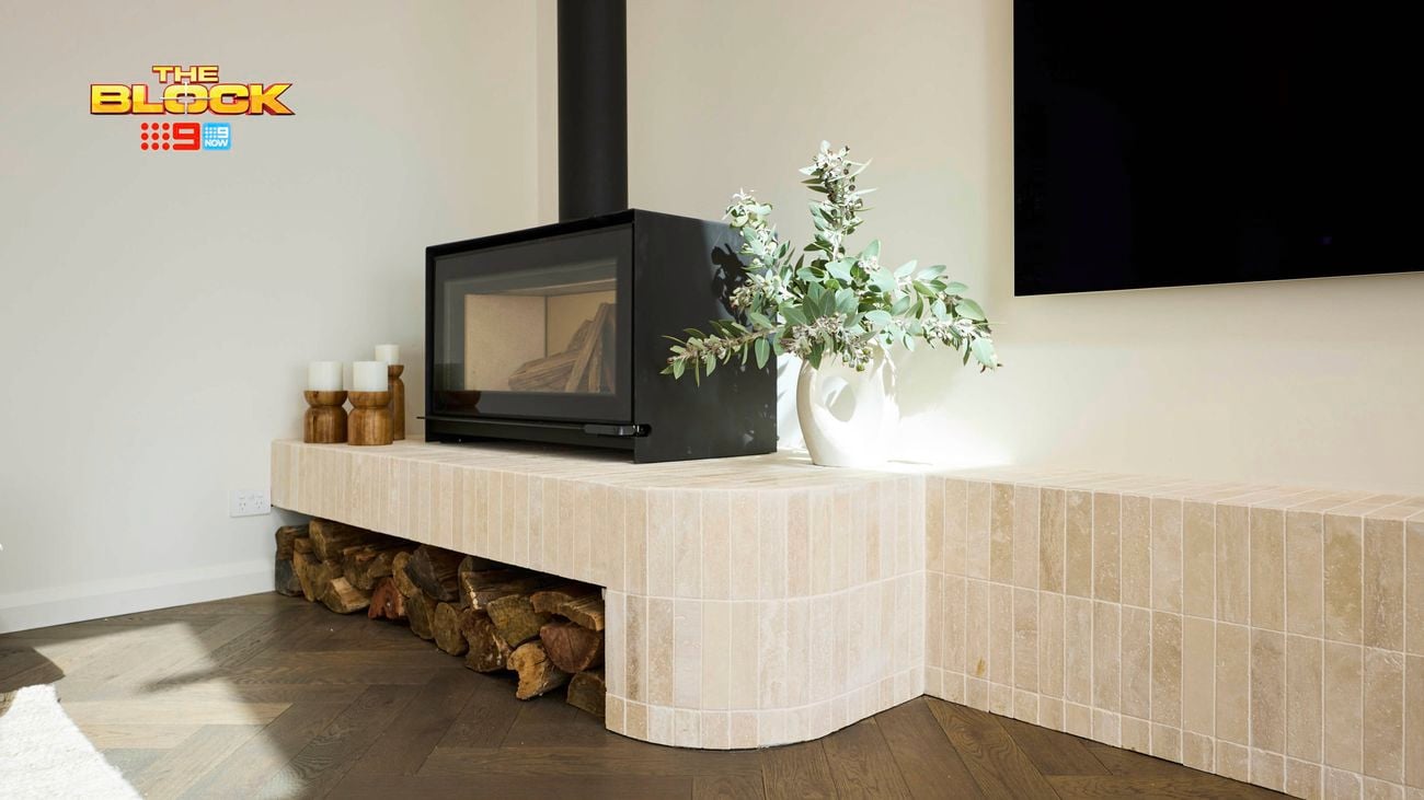 A modern living room corner featuring a black-framed fireplace built into a light-colored tiled structure that extends into a bench-like surface. Firewood is stored beneath the bench, and the top is decorated with a white vase of green foliage and candles. The floor has dark wood herringbone patterning, and a large black screen is partially visible on the wall. 