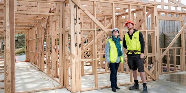 A woman and man, both wearing high-vis vests and hardhats, are looking up to inspect roofing trusses inside the frame of a house under construction.