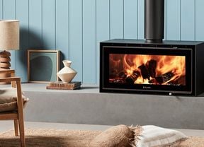 Black freestanding wood heater with burning logs against a light blue wall, next to a log holder with a rug and cushion on the floor.